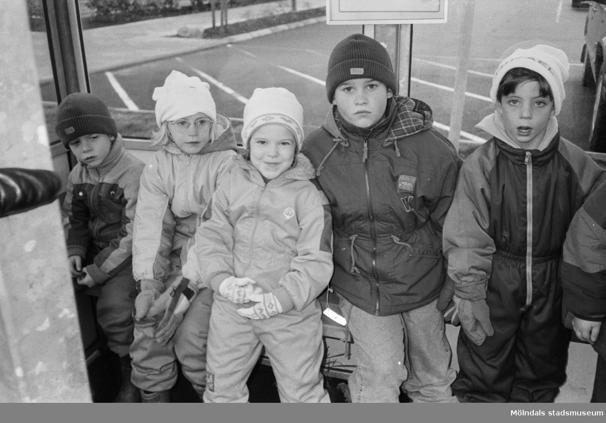 Fem barn, iklädda jackor, mössor och vantar, sitter/står i en busskur. I bakgrunden syns tomma parkeringsplatser. Utflykt med Katrinebergs daghem, 1992.