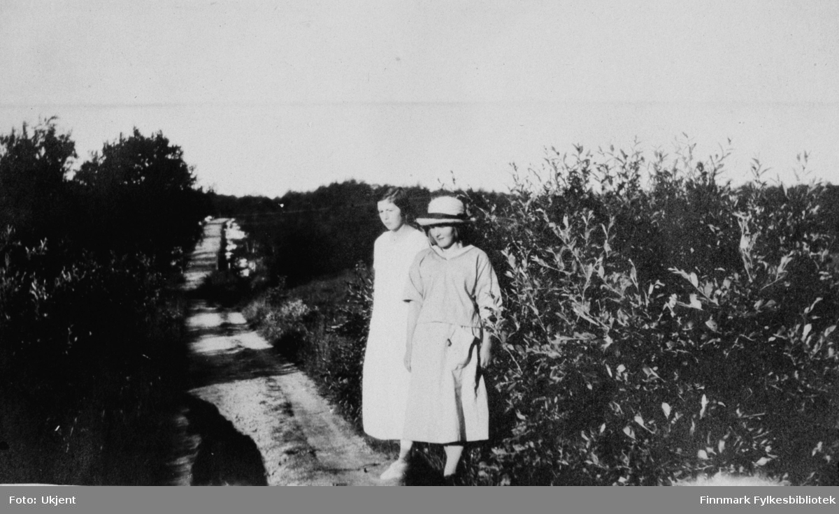 Bildet er tatt i Jarfjord sommeren 1924. Fra venstre: Elsa Bertelsen og Elin Anna (etternavn ukjent). De to jentene står på en grusvei som er omringet av planter, busker og trær. De er kledd i sommerkjoler, Elin har på seg en hatt.