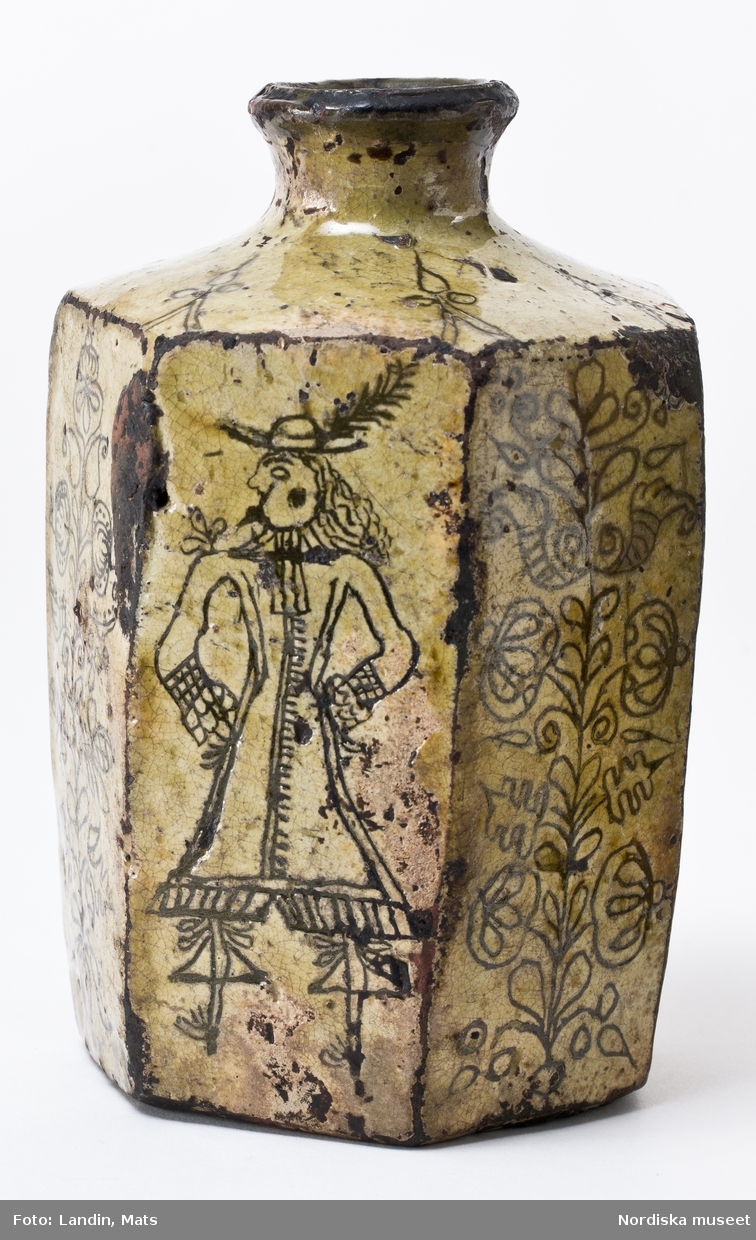 Sjusidig flaska av lergods, engoberad, med ristad dekor med man i 1600-talsdräkt och växtornament.
Se Bonge-Bergengren 1994, s 23.