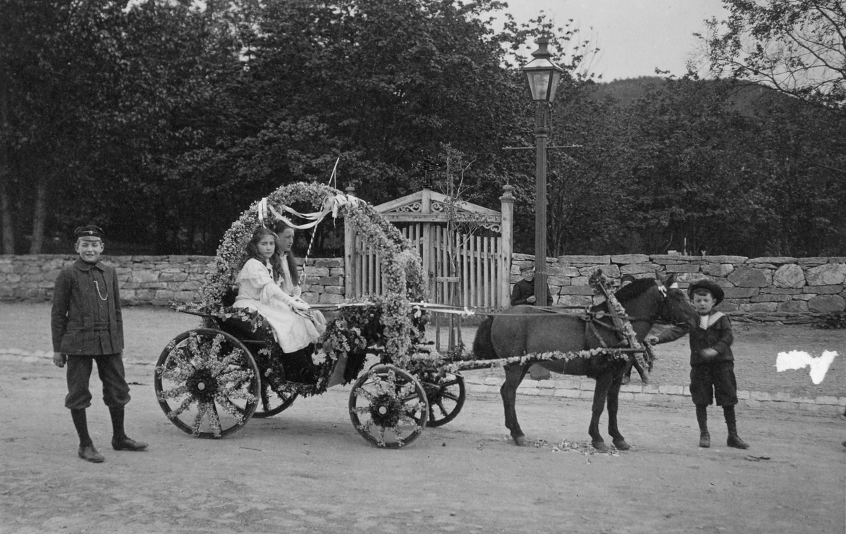 En ponni trekker en blomstersmykket vogn. Astrid Elsa, Marentius og Wilhelm August Thams i og rundt vogna.