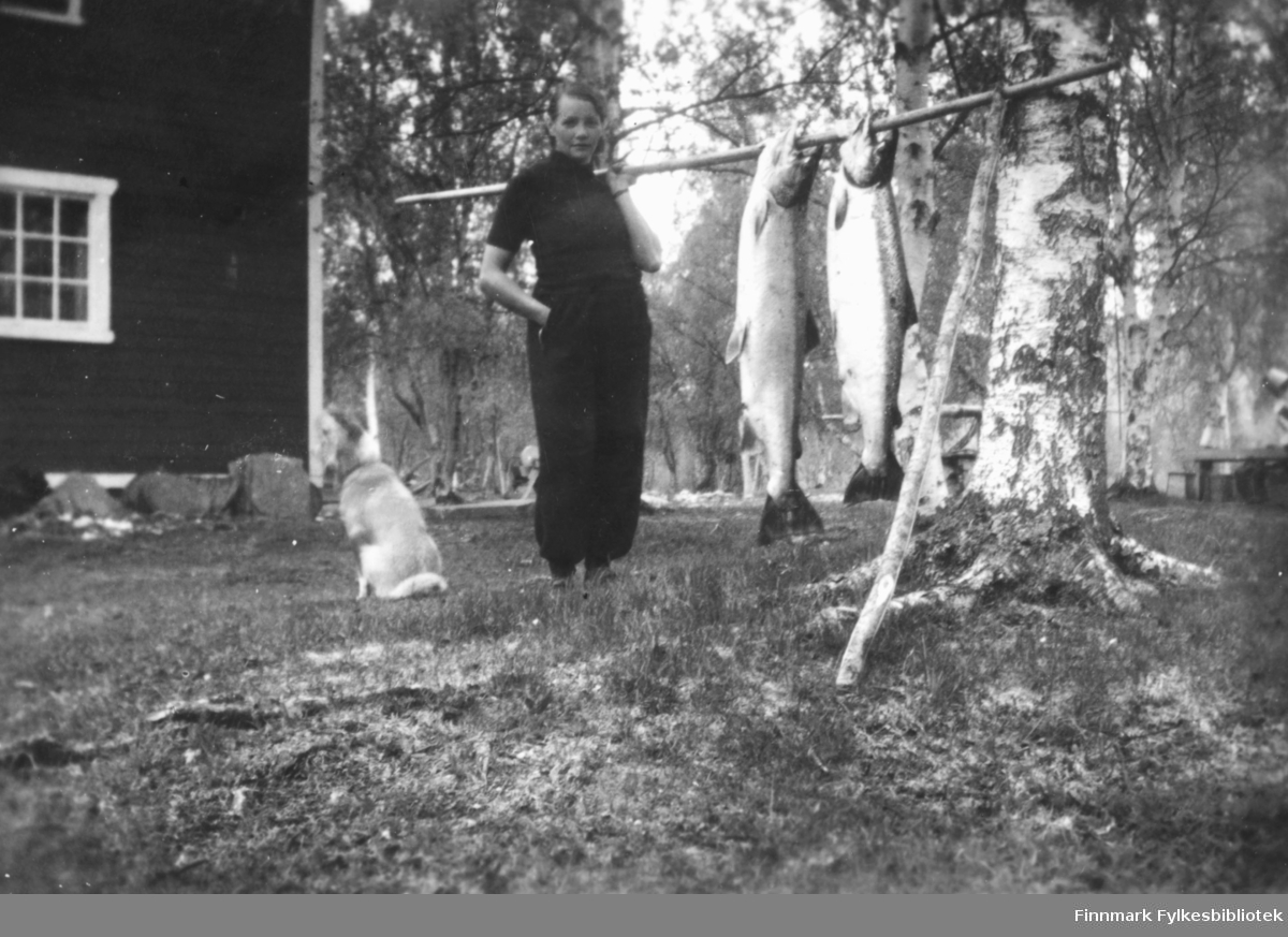 Magna Wisløff viser fram to store laks som henger fra en bærestokk. Til venstre for henne sitter en hund. I bakgrunnen ser vi et fjøs, gårdstun og trær. Ca.1930-40.