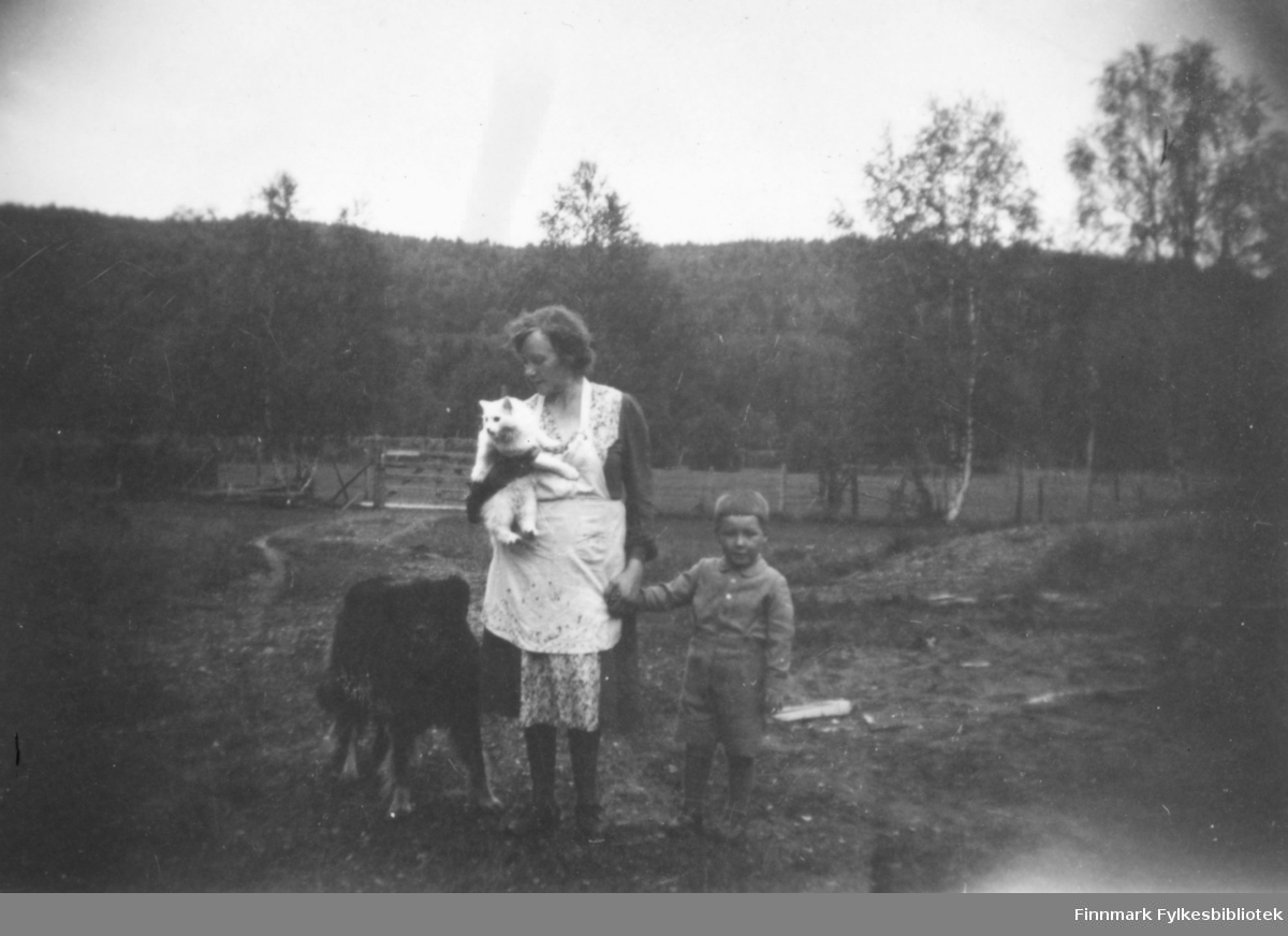Magna Wisløff leier sin sønn Ole Arve Wisløff i hånden. Hun holder familiens katt og til venstre for henne står Kristian Wisløffs hund. Bak dem ser vi et gjerde og et jorde. I bakgrunnen tett skog. Ca.1945-50.