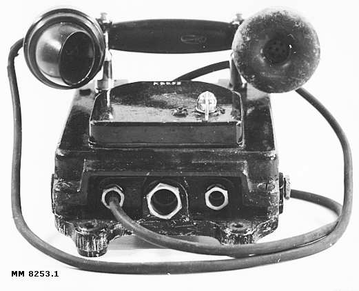 Bojtelefon av väggmodell, vattentät och svartmålad. L.M. Ericssons tillverkning. innesluten i ett hölje av gjutjärn. Utanpå telefonen ett fäste för handmikrotelefon.