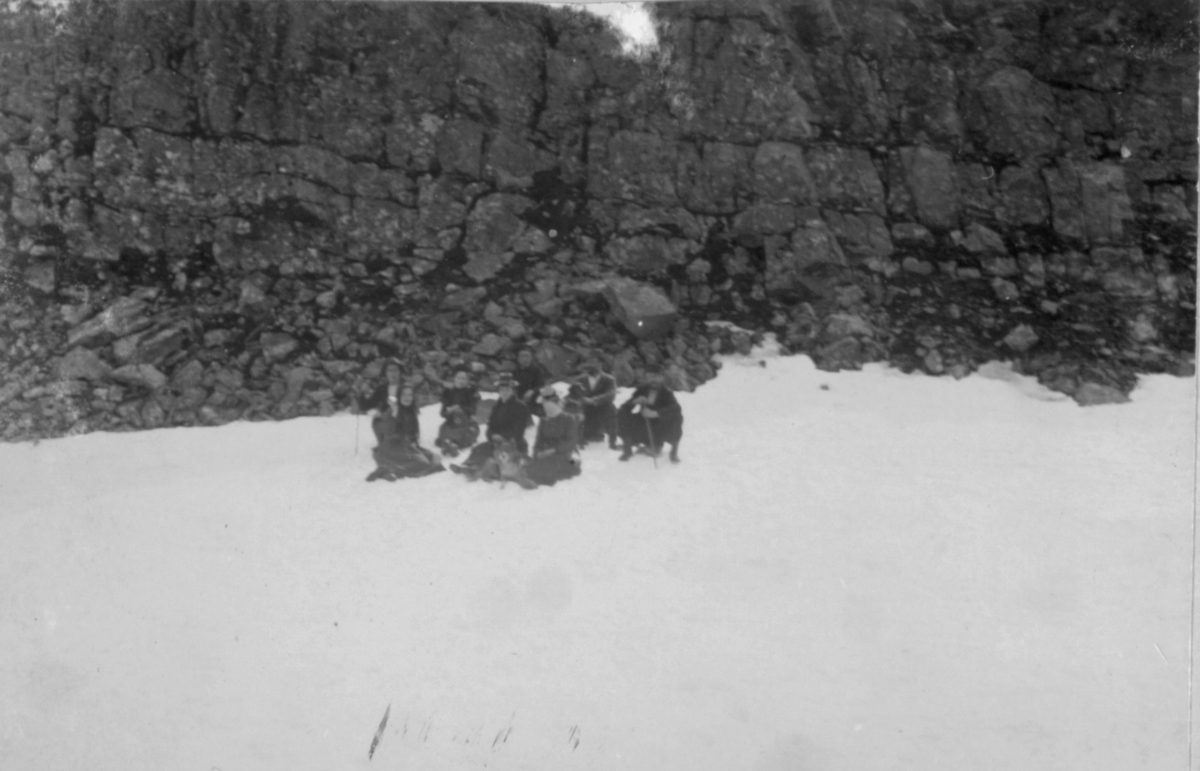Damer og menn sitter i snøen ved en steinur.