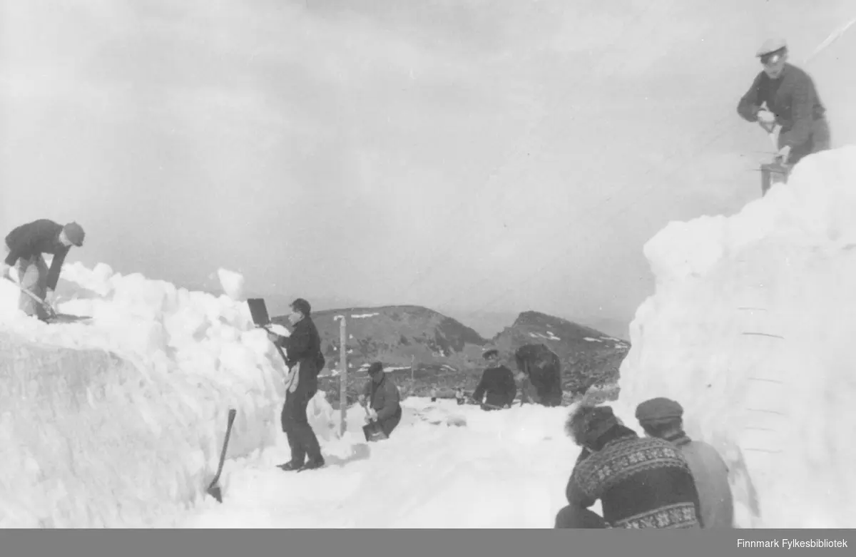 'Veirydding mellom Hasvåg og Hasvik i mai 1946. Veien måtte ryddes forå få fram materialer til Hasvik, hvor der ikke var kai." I bildet ser vi flere menn med spader og håndkraft rydder snø av veien.