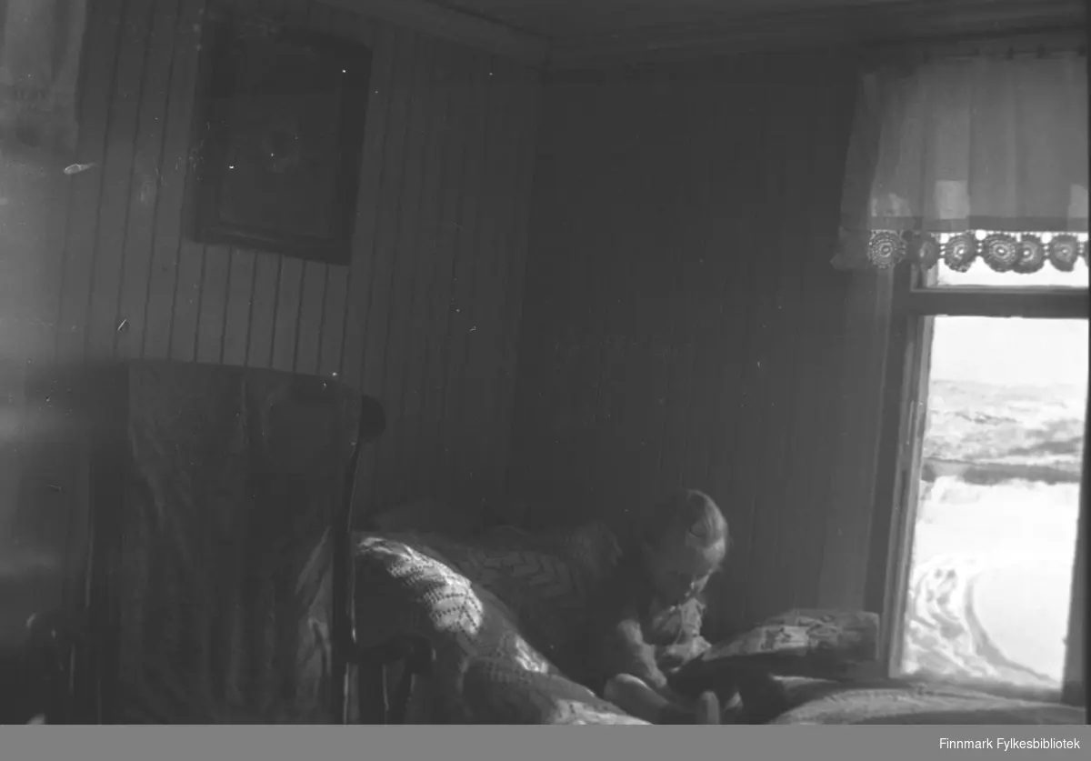 Herlaug Mikkola leker på senga inne på Mikkelsnes. Hun sitter på et fint heklet sengeteppe, og i vinduet er det gardiner med en vakker heklet kant