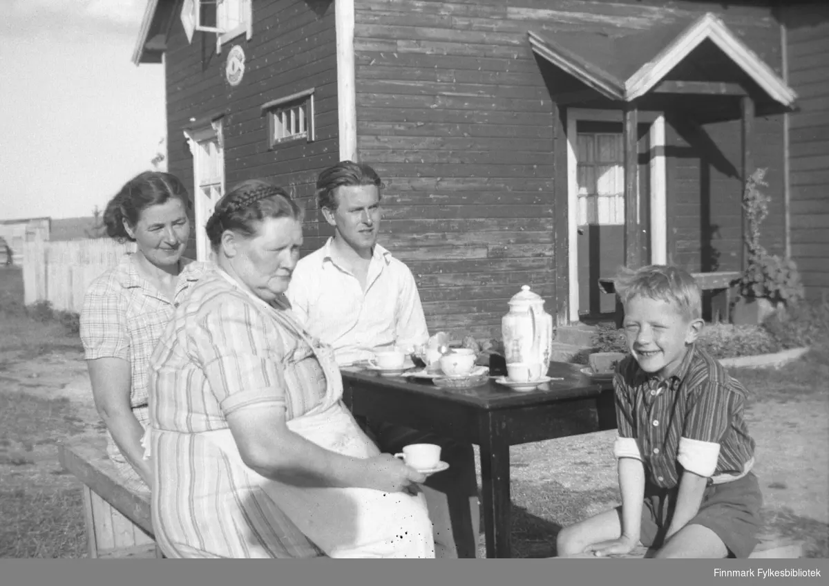 Ukjent par på besøk på Mikkelsnes, ca. 1947-1949. Her sitter de ute og drikker kaffe. Merk talestasjon-skiltet på veggen på våningshuset i bakgrunnen. Kathinka Mikkola sitter med kaffekoppen i handa. Barnebarnet hennes, Per Olsen, sitter til høyre og smiler mot fotografen