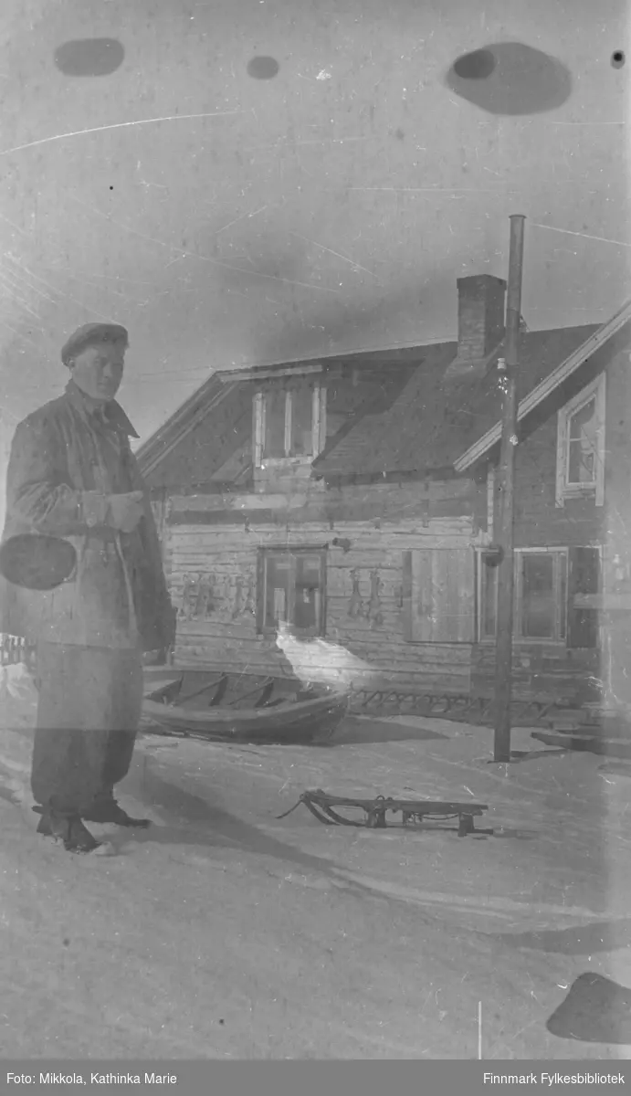 En ung mann i vinterklær, Andreas Mikkola bak våningshuset på Mikkelsnes. Han står ved en liten kjelke. I bakgrunnen en robåt trukket opp ved siden av huset. Det er sperret opp små skinn på husveggen - hareskinn?