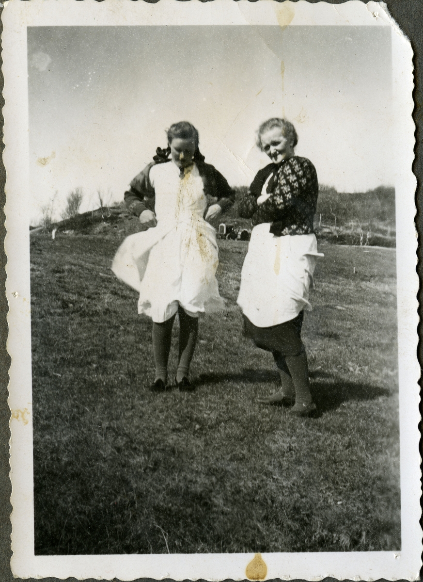 Fra venstre: Åse Reeberg og mor Anna Reeberg. Anna er kledd i strikket jakke med mønster, forkle og kjole. Åses kjole blir tatt av vinden. Hun har fletter og sløyfer i håret. I bakgrunnen, mellom damene, kan man se en traktor.