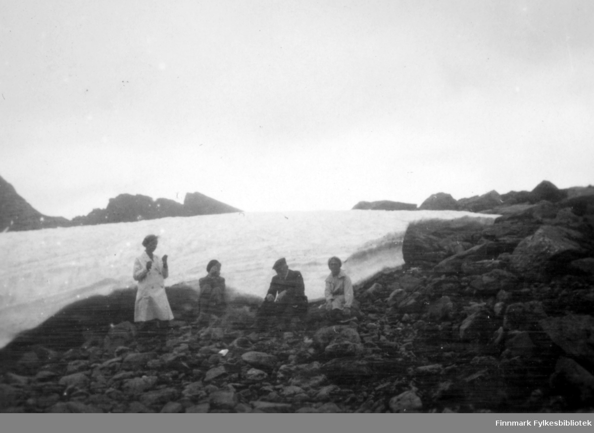 Fire personer sitter ved fjæra. Snøen er ikke helt smeltet, vi ser rester av vinterens snø i bakgrunnen. Damen til venstre på bildet har en lys kjole med korte ermer på seg. Ved siden av henne sitter en dame i ganske mørke klær. Mannen har en mørk dress og sixpence på hodet. Damen til høyre på bildet har et lyst skjørt på seg. Stedet er i Bergsfjord i Loppa rundt 1920-1930.