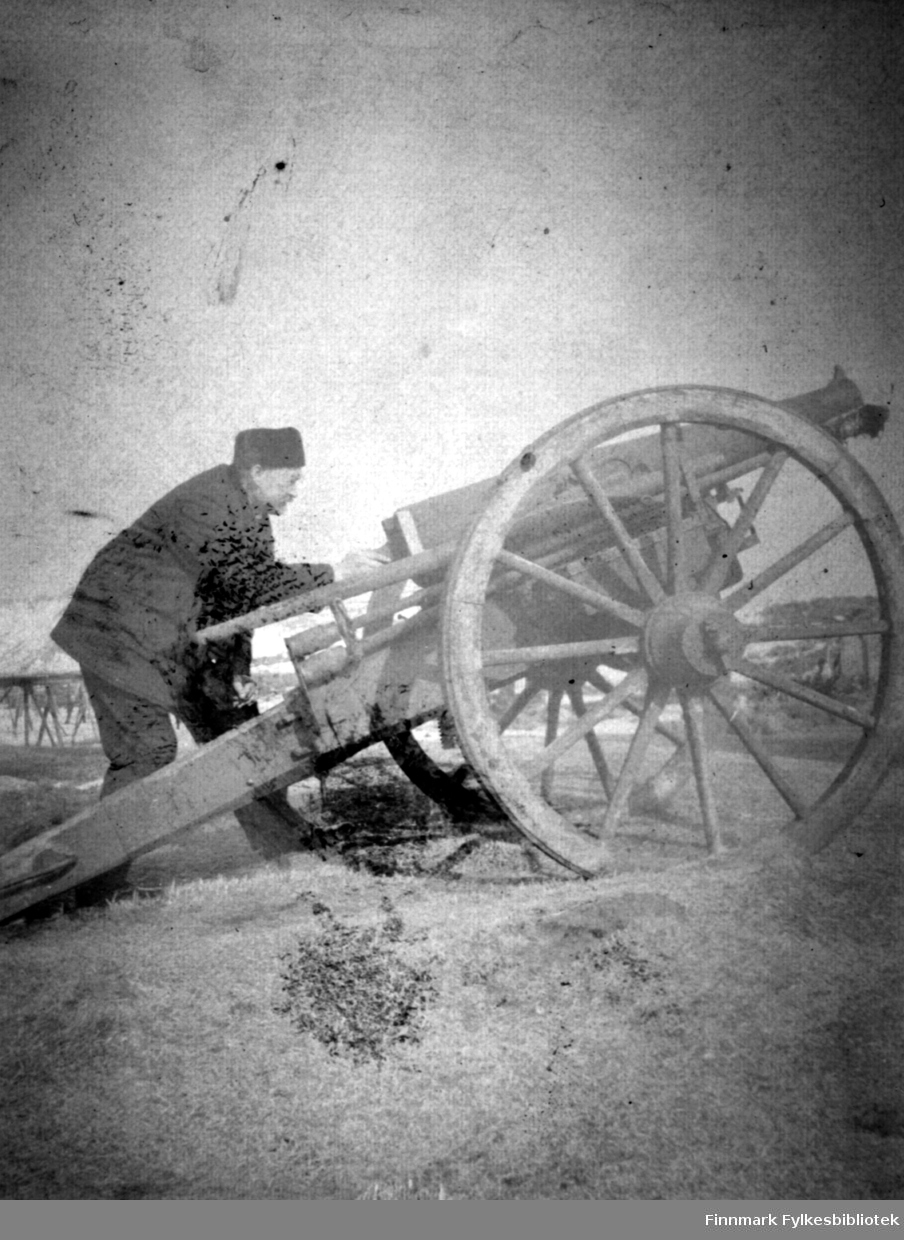 En mann i mørke klær og lue på hodet står bøyd bak en kanon, muligens en Krupp feltkanon. Kanonen har trestativ med lang 'hale' og store trehjul. Det er gress og lyng på bakken og noen snøflekker kan ses i terrenget i bakgrunnen.