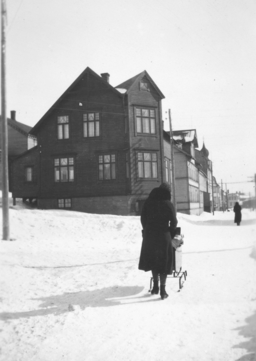 Vinteren 1934 i Havnegaten. Bildet et tatt fra omtrent der avisa 'Finnmarken' har sine lokaler. Rett frem ligger Sellstrømgården som Hauge familien flyttet til. Alfrida Hauge går på veien, med sønnen Tor Hauge i barnevogna.
