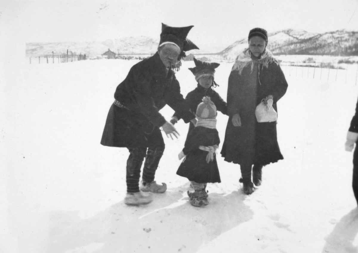 En mann, en kvinne, og to barn kledt i kofter, og samiske hodeplagg. På bena har de skaller. Det er vinterstid, og det ligger snø på bakken. Bildet er muligens tatt i Aleknjarg området