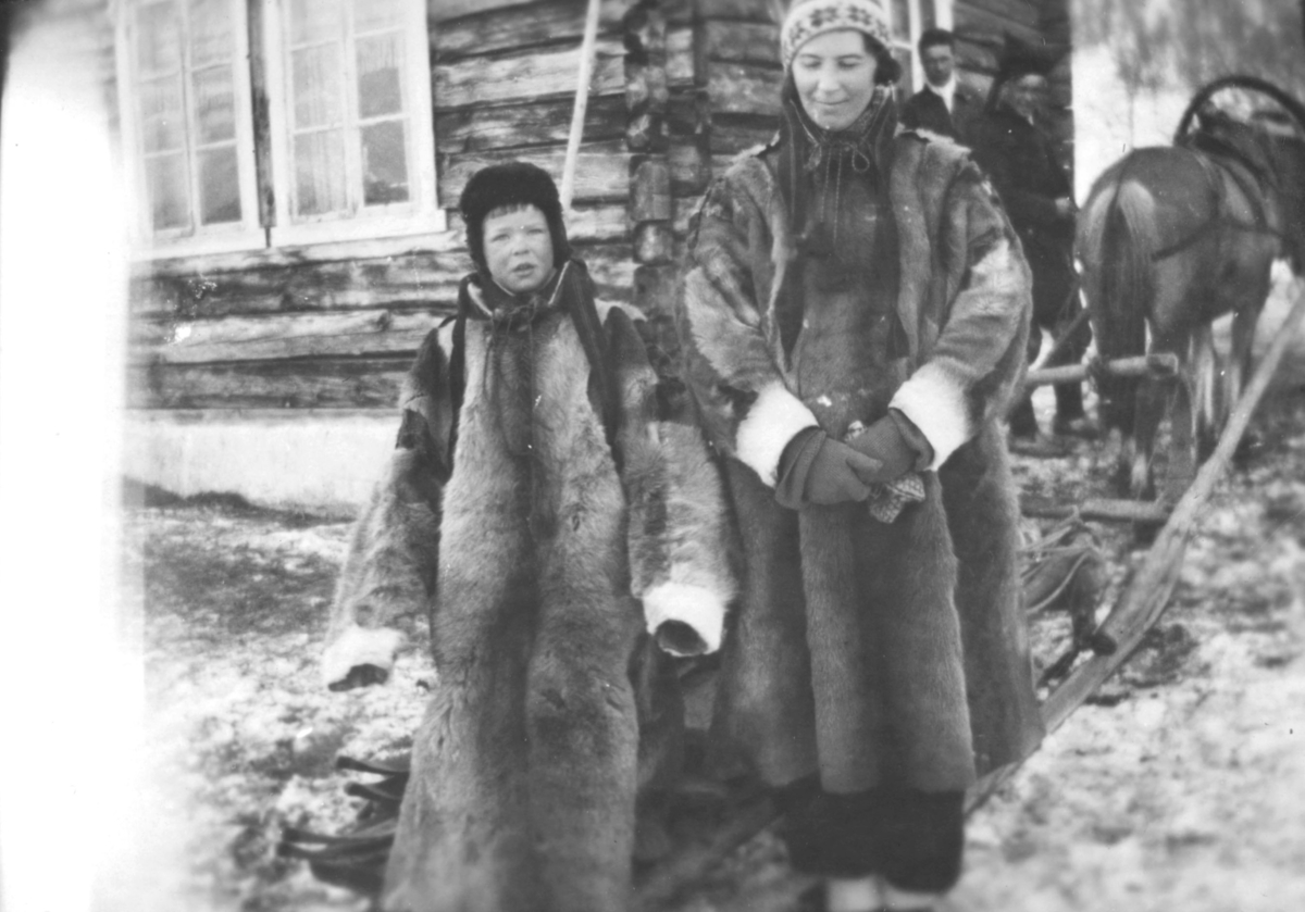 Tor Hauge, og moren Alfrida Hauge f. Amundsen, i lånte pesker. De står foran en tømmerbygning. I bakgrunnen står to menn, ved siden av en hest. Hesten er spent foran en slede. Det ligger snø på bakken