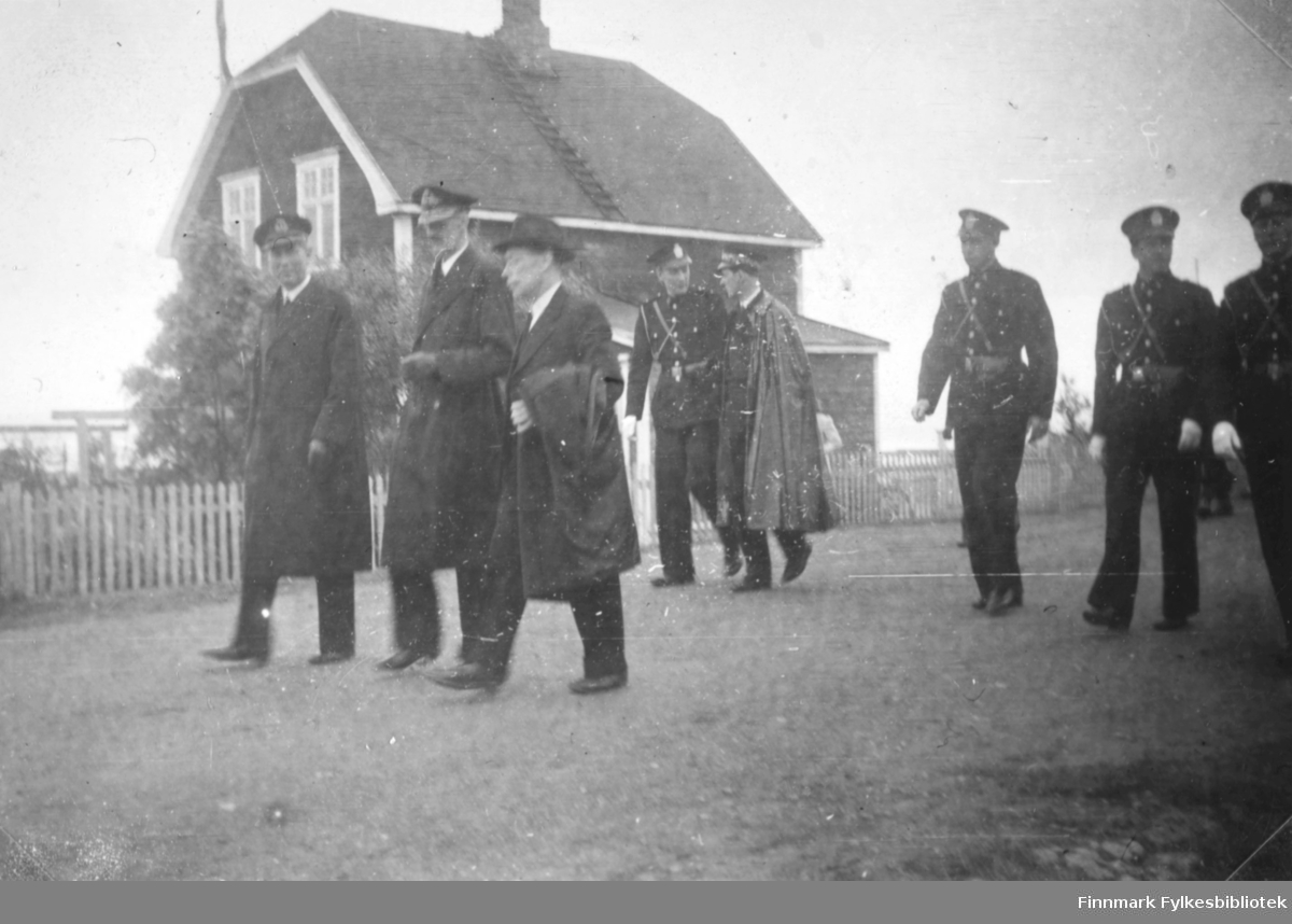 Kong Haakon VII besøker Vadsø den 12.07.1946. Her er kongen på vei sammen med fylkesmann Hans Julius Gabrielsen gjennom Vadsøs gater. Huset i bakgrunnen tilhørte Georg Evanger. En av politimennene er visstnok Gullik Iversen.
