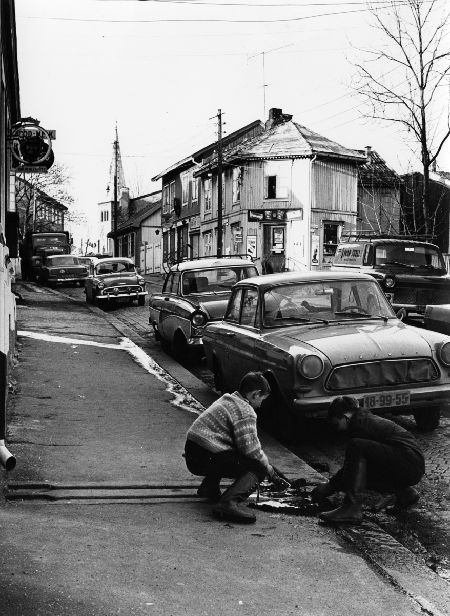 Foto av to gutter som leker på fortauet i Normannsgata på Kampen. Kirken ses bak den gamle trehusbebyggelsen. Biler står parkert i gata. .