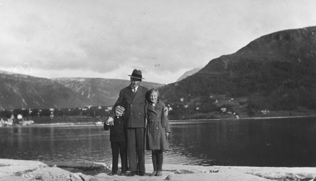 Bildet er tatt i Tromsø. Fra venstre Rakel Lein, lærer Ødejord og Lars Ødejord. I bakgrunnen ser vi sjø og fjell. Mannen er i mørk dress og hatt. Den ene amen holder han rundt jenta som har kåpe med belte på. De andre armen holder han rundt en gutten som har mørke klær på.