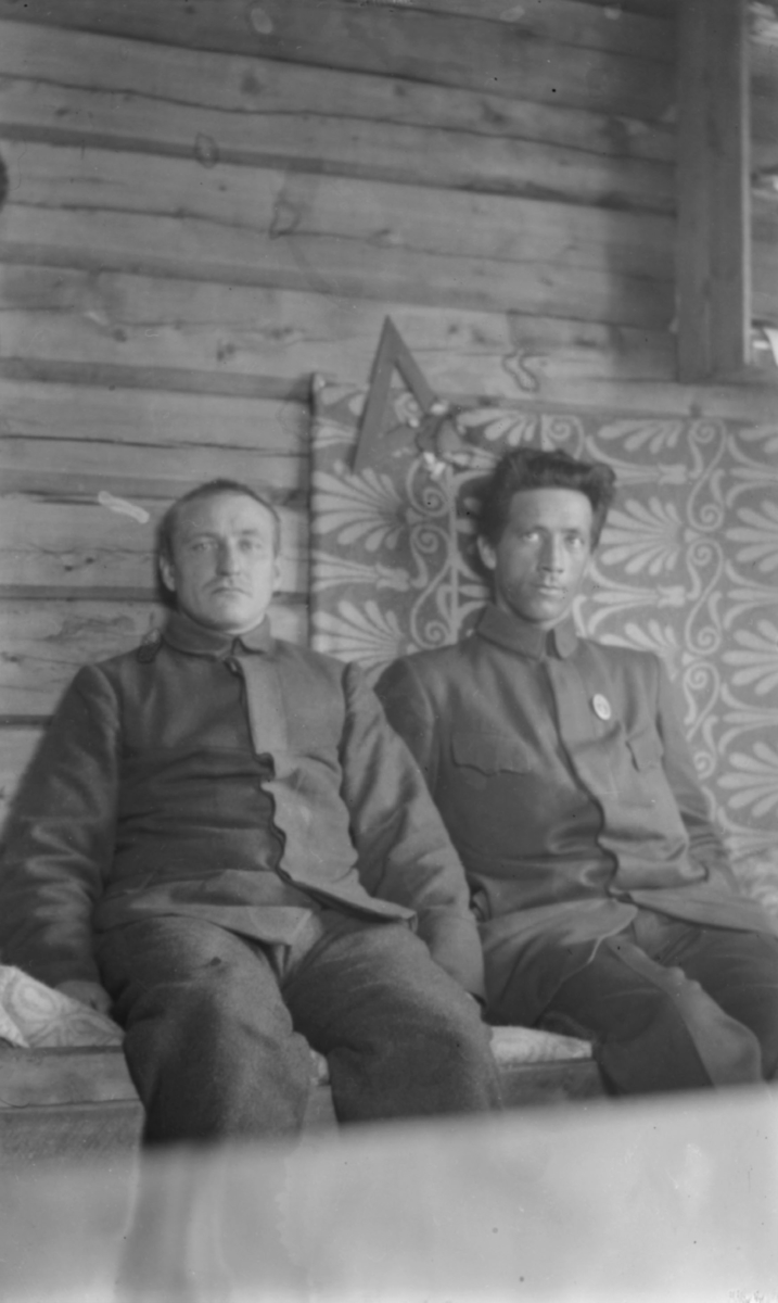To menn sittende inne ved en tømmervegg - de er kledd i uniformer. Dette kan ære interiør fra en kaserne, jfr. bildets påskrift. Bak mennenes rygg et tekstil i vakkert liljemønster