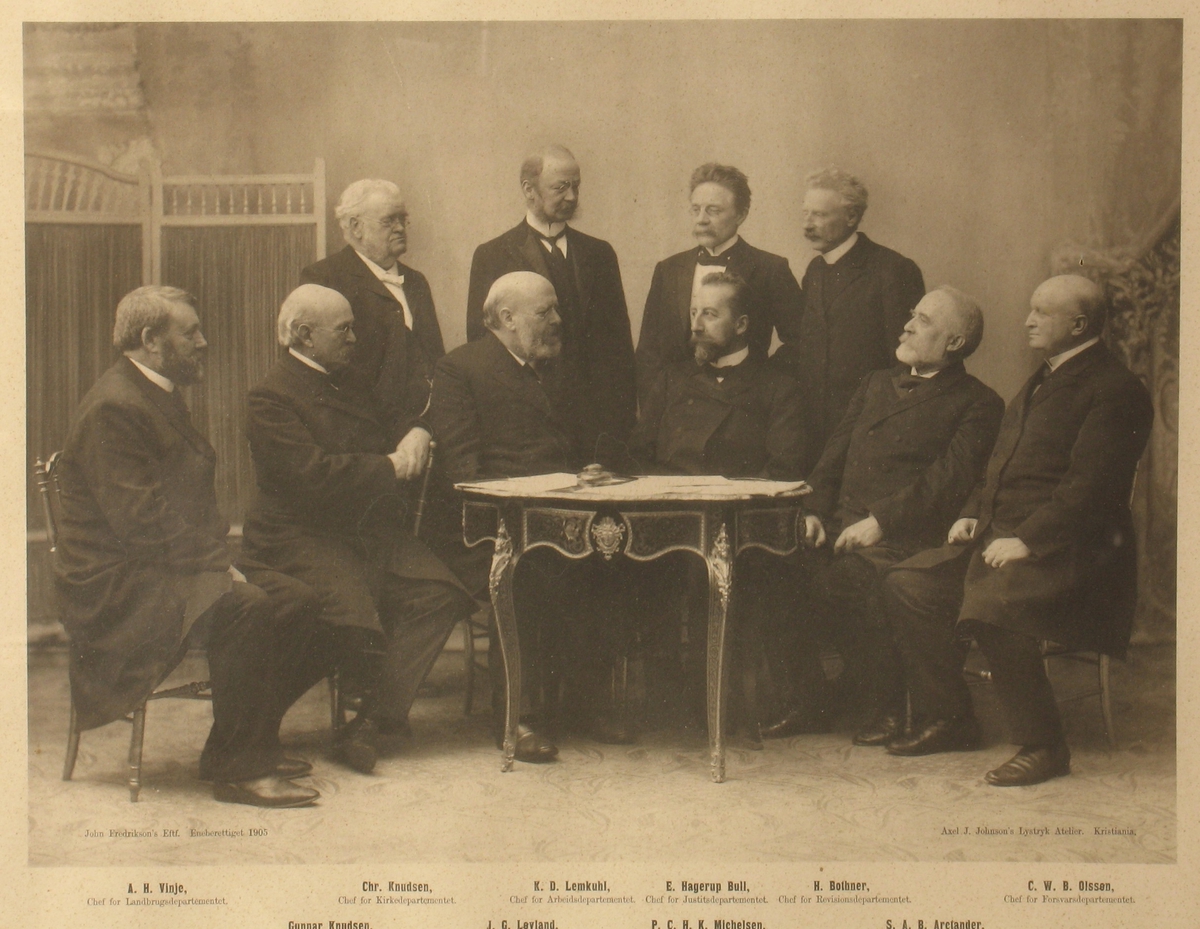 Gruppeportrett av den norske regjering i 1905,  10 menn samlet rundt et bord. 6 sitter på stoler, 4 stående bak. De 5 på høyre side ser mot venstre, og vise versa. Navnene er trykt i bildets nedkant.