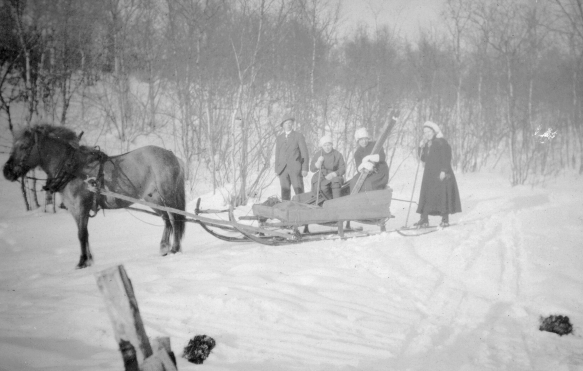 Hest og slede med mennesker. På tur fra Jarfjord til Knes. Flere kvinner og menn er kledd i vinterklær. En kvinne som sitter i sleden holder på et par ski, og kvinnen t.h for henne står på ski.