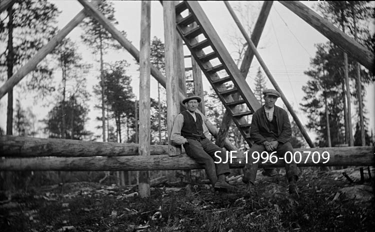 To menn sittende ved foten av brannvakttårnet på Haukenesfjellet i Rømskog i Østfold.  Denne lokaliteten har hatt tre brannvakttårn.  Det første ble reist av Østfold Skogselskap i 1909 og inngikk da i et nettverk med Vedøfse i Aremark, Høyås i Asak (Halden) og Linnekleppen mellom Aremark og Rakkestad.  Det første tårnet råtnet og ble i 1928 erstattet av et nytt tårn.  Tårn nummer tre ble reist i 1953.  De to siste tårnene ble reist i samarbeid mellom Østfold Skogselskap og forsikringsselskapet Skogbrand.  Skogbrannvaktholdet på Haukenesfjellet ble avviklet i 1976.  Etter den tid har lokale flyentusiaster fått betaling for å gjøre rekognoseringsturer.  Det siste tårnet på Haukenesfjellet ble i 2007-08 overtatt av Den norske Turistforenings avdeling i Østfold.  Turistforeningen vil restaurere tårnet med sikte på å innrede overnattingsrom for vandrere.  Om lag samtidig har Norsk Skogmuseum bygd en fullskalakopi av dette tårnet på sitt uteområde med støtte fra Skogbrand. 

Fotografiet viser antakelig det andre tårnet på Haukenesfjellet (1928-1952).  Det ble reist av tårnbyggeren Anton Lillevold (1893-1978) fra Furnes i Hedmark og hans mannskap.  Tårnet var en slags standardmodell, cirka 10 meter høgt og sammensatt av om lag 65 tømmerstokker.  Hvem de to avbildete karene er har vi foreløpig ingen informasjon om.