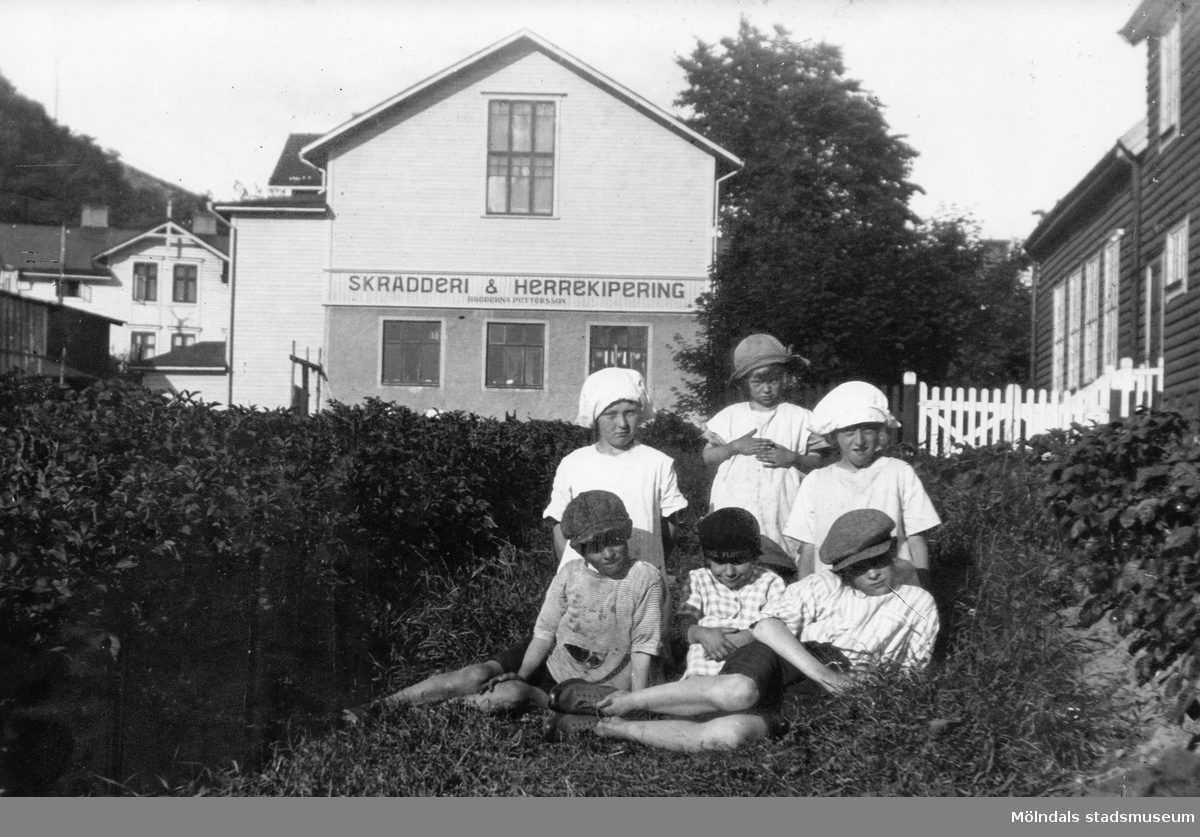 En samling barn sittandes för fotografering framför Bröderna Pettersson Skrädderi & Herrekipering vid Kvarnbygatan i Mölndal på 1910- eller 1920-talet. I bakgrunden ses hus vid Trädgårdsgatan, i mitten gamla Folkets hus och till höger Frälsningsarmén.