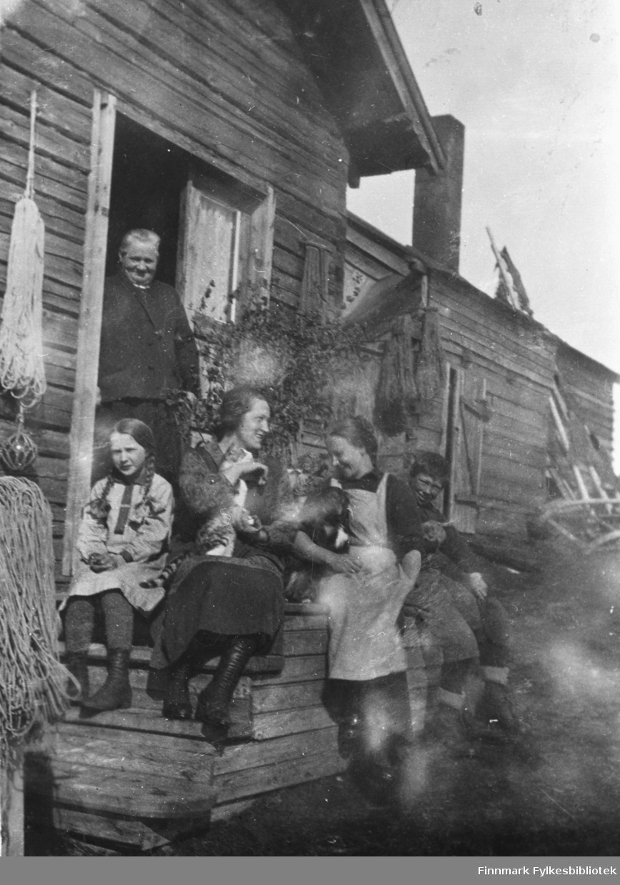 Fru Ballo står ved dørren i familiens hus i Vestre Jakobselv. På trappa foran hun sitter en jente med fletter, to unge kvinner og en gutt. Den ene kvinnen har en katt i fanget og den andre en hund i armkroken. Det står bjørkekvister ved dørrn, en skikk fra Finland på st.Hans-feiring.