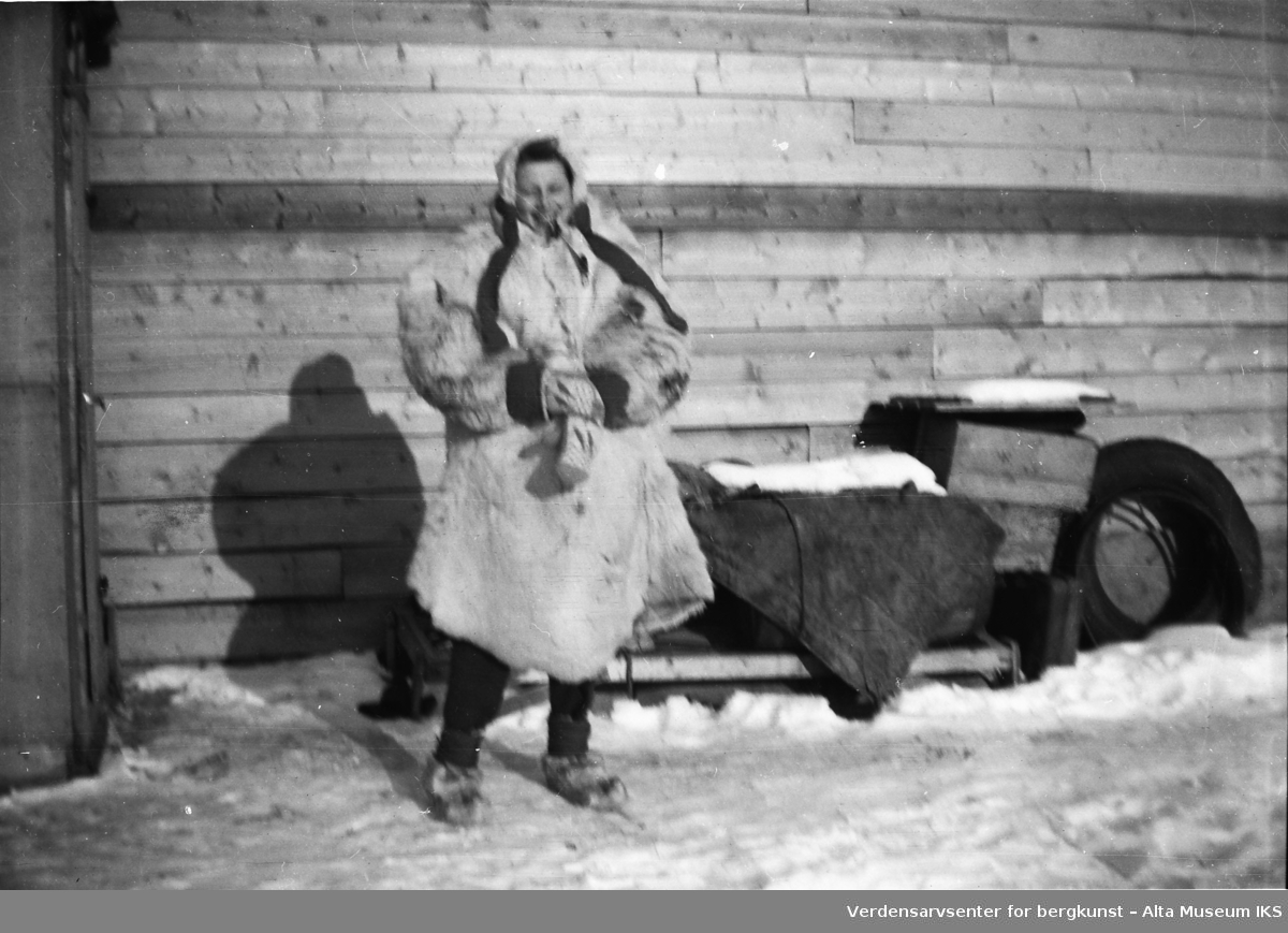Kvinne iført tjukke vinterklær av pels står foran en husvegg.