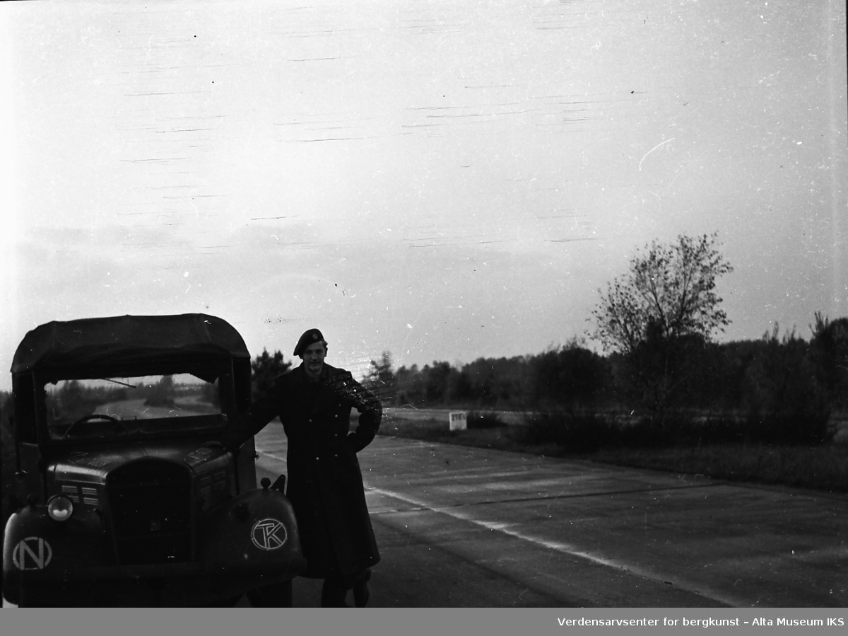 En ung mann i militæruniform og en berret lener seg mot en militær bil på en tom motorvei.