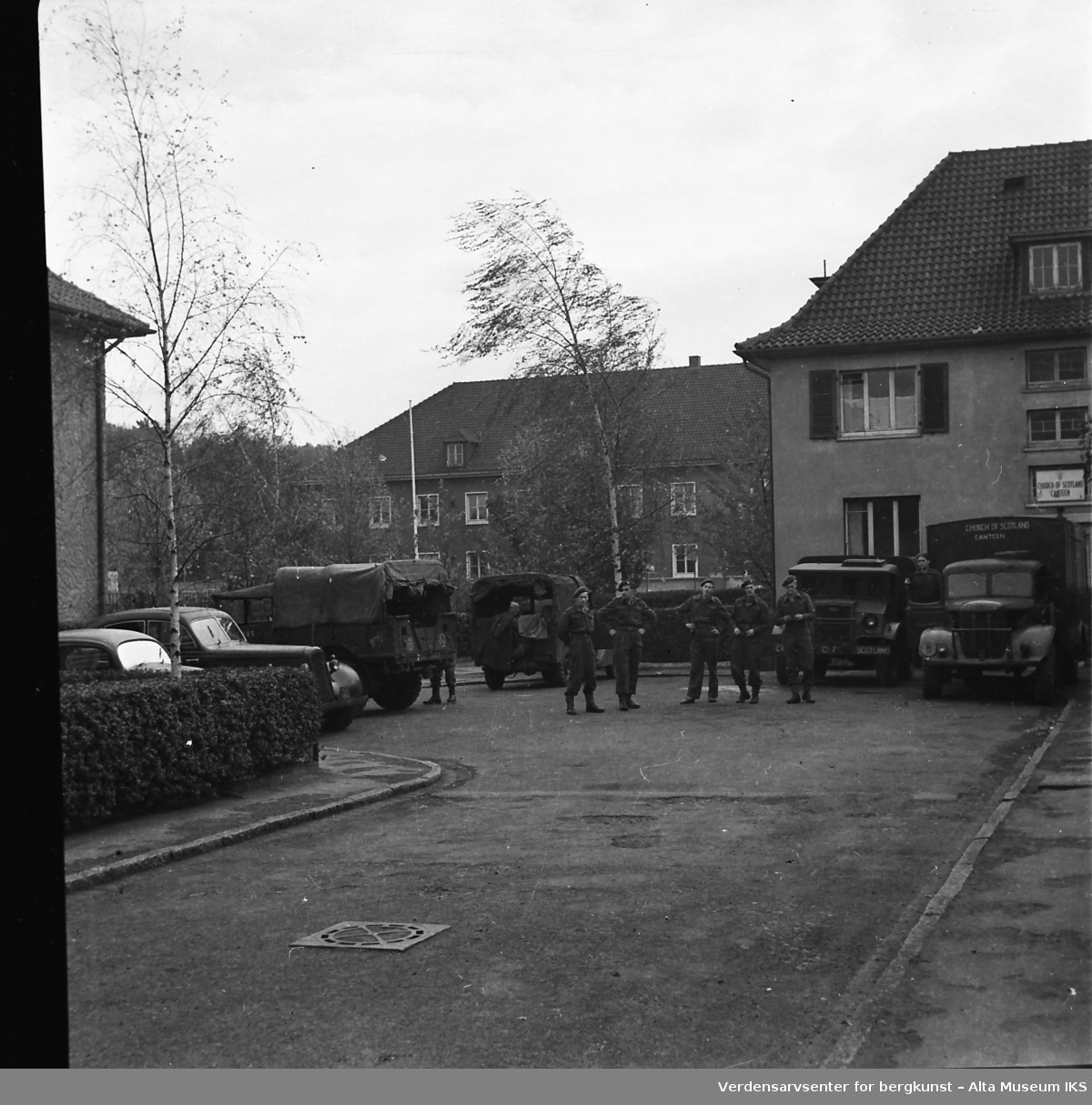 Fem uniformerte menn står foran militære lastebiler i en gate.