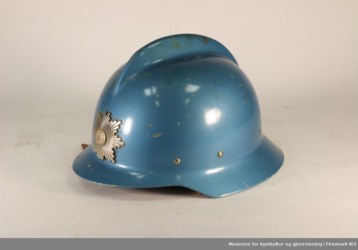 Blå brannhjelm av metall med hanekam på toppen. Foran er det brannvesenets emblem. Innerfor av hjelmen er av skinn. Foret kan reguleres med snor.