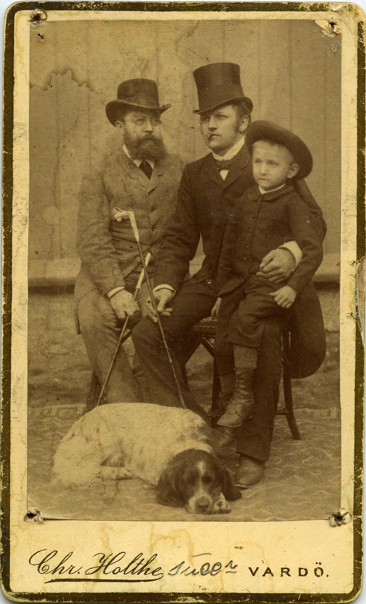 To ukjente menn og en liten gutt. Gutten sitter på fanget til den yngste mannen, en hund ligger ved siden føttene til avbildete personer. Den eldre mannen med skjegg kan være Bersvend Thoresen.