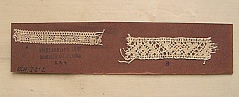 På en bit brun pappkartong är två små, vita, knypplade spetsprov uppklistrade.
A. 8 x 1 cmB. 8 x 1,8 cm Hjärta,halv trissa, ljusgullsskack