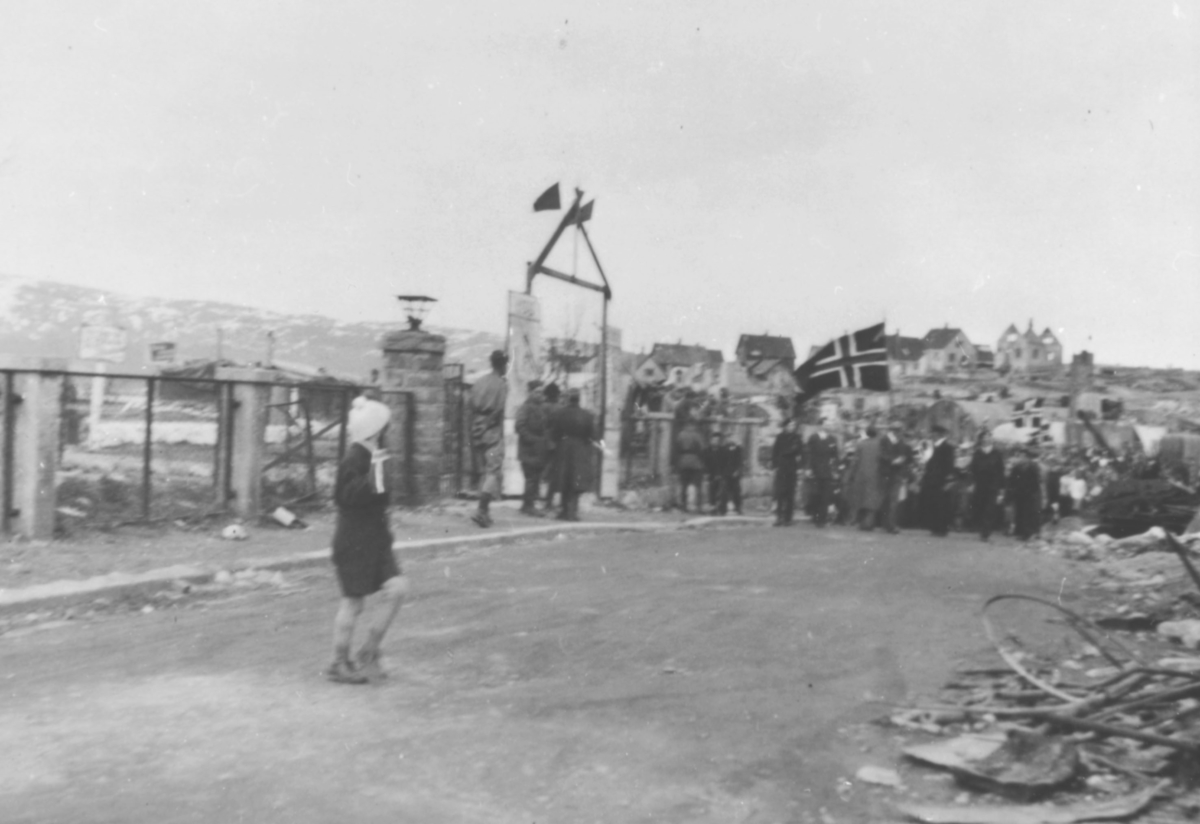 17.mai i Kirkenes, 1945. Tog med et par flagg. I forgrunnen et barn. I bakgrunnen boliger, delvis ødelagte.