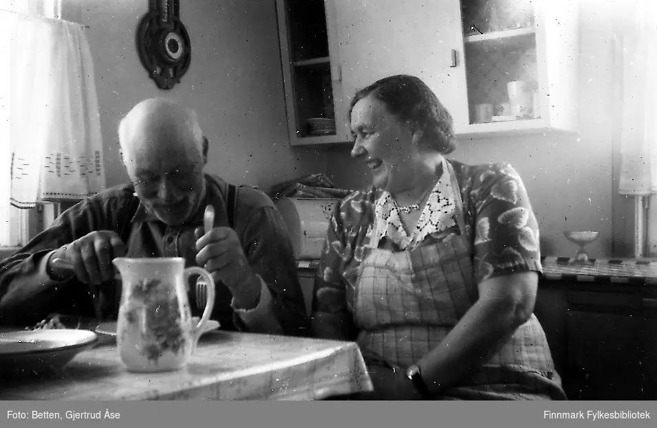 En eldre ektepar, Edvard og Karoline Johnsen, sitter i kjøkkenet ved kjøkkenbordet. Edvard sipser middag, Det står en porselenkanne på bordet. Bordet er plasert ved vinduet, litt av gardinene vises. Bakerst kjøkkenbenken og kjøkkenskap.