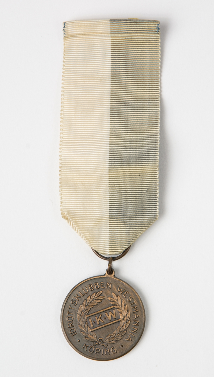 Medalj i brons från F1 Medalj med band. Medaljens framsida har en lövkrans och innanför den initialerna I.K.W. Runt kransen lyder texten: IDROTTSKLUBBEN WESTMANNIA KÖPING. Baksidan har en liknande krans. Medaljen är fästad i ett blått/vitt band.