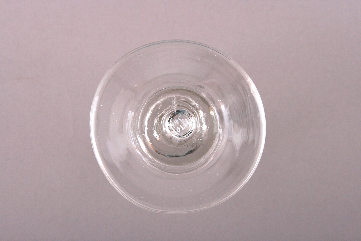 Lite glass med inngravert monogram; MTW el. MJW. Monogrammet er omkranset av en bladkrans. Over er det en krone med 5 tagger. Monogrammet er på begge sider av glasset. Det er et lite skår i drikkekanten.