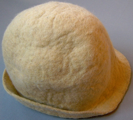 Tovad hatt i naturvit ull. Rund kulle med smalt brätte. Hatten är något stel och gulnad.