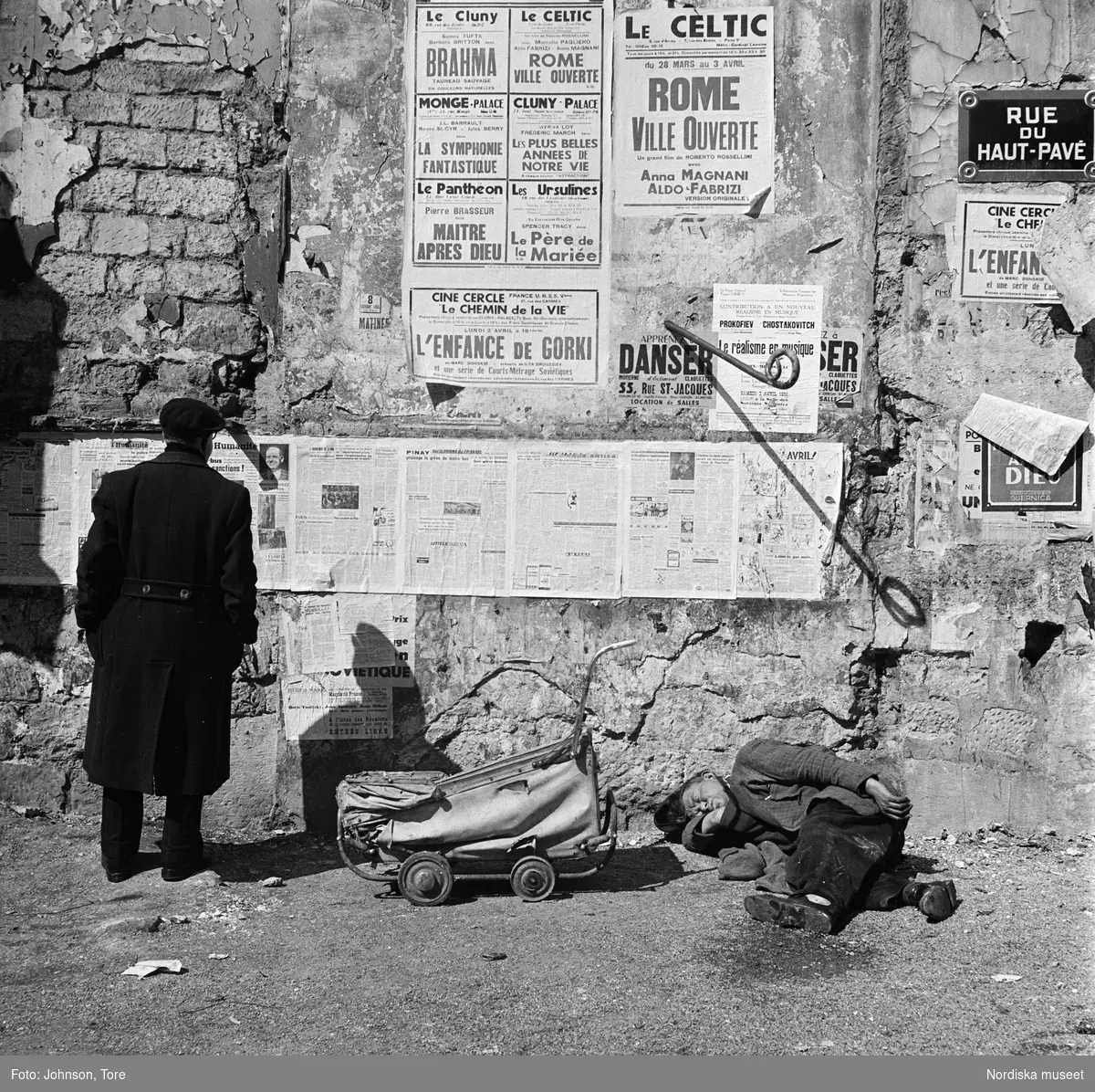 Paris, Rue du Haut-Pavé. Två män, en stående läsandes en väggtidning, den andra sovande. En dockvagn i mitten.