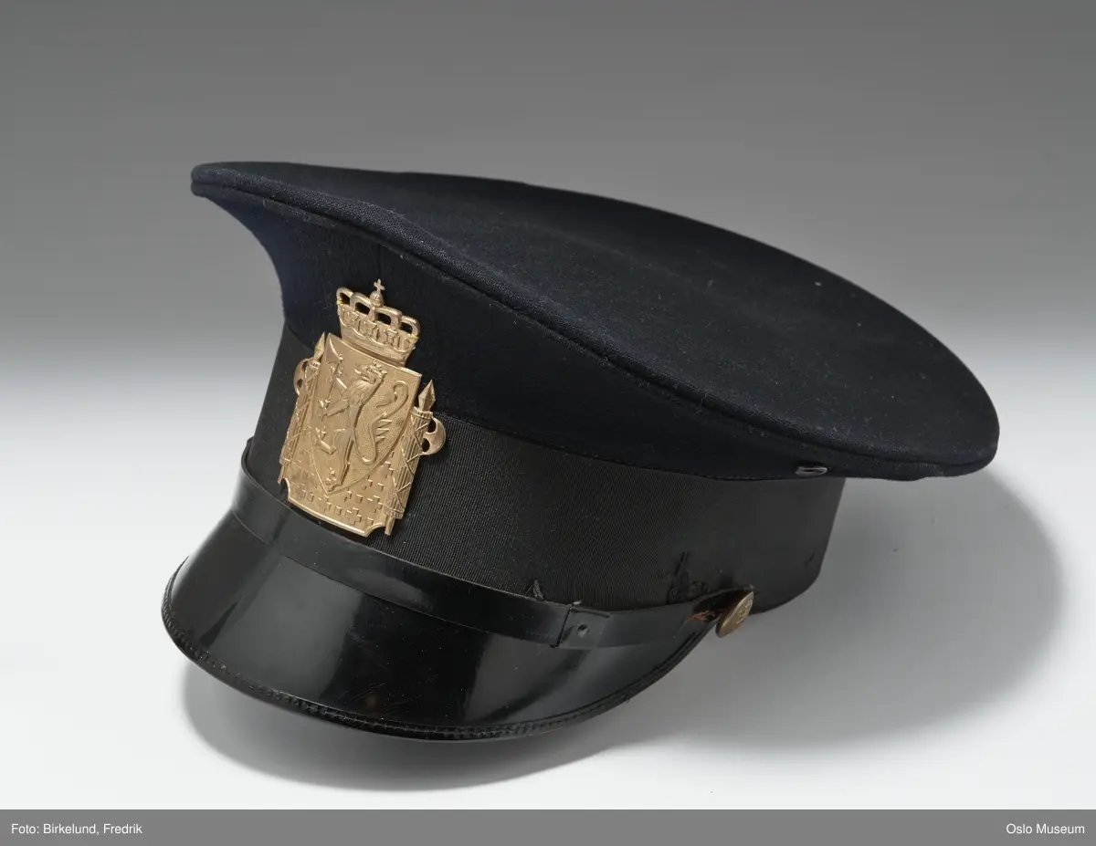 a) Jakken: Kraven oppstående, lukning med to metallhemper og kramper. Lukning foran med seks støpte, hule gultmetalls knapper med den norske løve. To brystlommer med klaffer. Lange ermer med påsydd kant. Fôr av stripet, sort silke, foran underforet med strie, ermene fôret med beige silke. På kraven foran festet to støpte gultmetalls løver.  b) Luen: Skyggelue. Pullen av mørkeblått klede med oval, flat, oppsvunget overside, sidene rette, høye, trukket med ripsbånd. Skjerm og stormrem av sort lakk. Svetterem av beige kunstlær, i pullen firkant av beige voksduk. Remmen festet med to knapper som jakkens, i fronten festet presset gultmetalls kronet skilt: Rektangel med den norske løve, på sidene fasces, bunnen med kors.