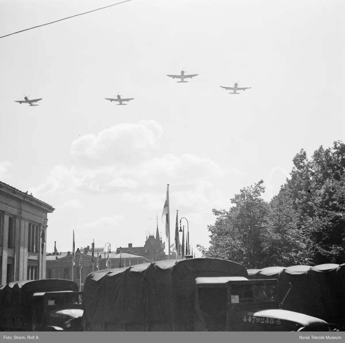 Opptog, USAs nasjonaldag, militærfly flyr over militære lastebiler ved Universitetet, gata er pyntet med norske, amerikanske og andre nasjonaliteters flagg, Oslo, 4. juli 1945.