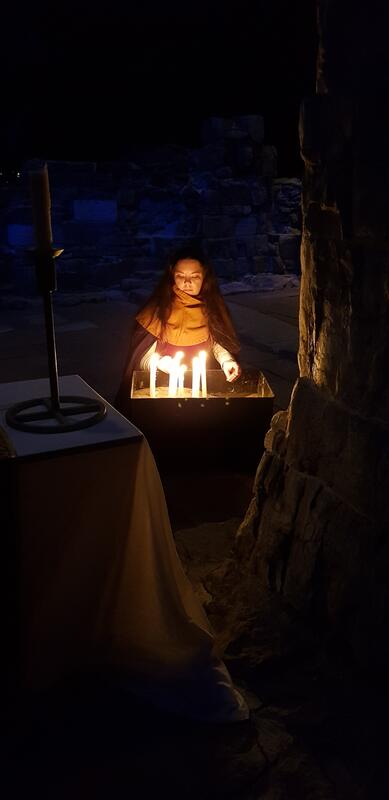 En middelalderkledd ung kvinne tenner levende lys under ruinbuene i Hamardomen.
