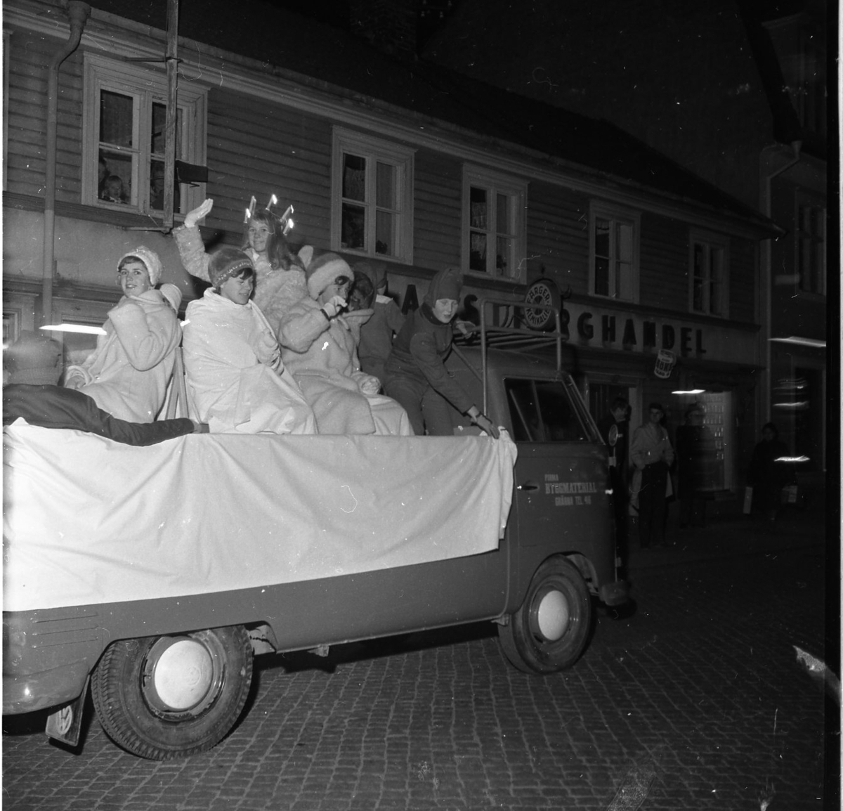Gränna scoutkårs lucia med tärnor sitter varmt klädda på en VW-pickup på Brahegatan i Gränna. Lucian, Marita Wallmo, vinkar mot fotografen. VW-pickupen är märkt "Firma Byggmaterial"