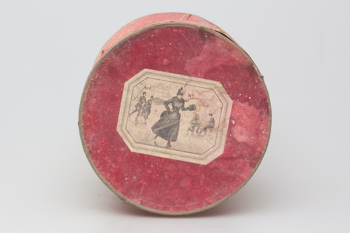 En rund, rødfarget eske med lokk som har inneholdt en muffe. På lokket er det en etikett med en tegning av unge damer og herrer som går på skøyter.