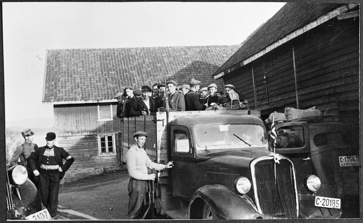 Fra mai-dagene 1945. Finn Gjestvang og Håkon Flenes ved bil med reg.nr. E-3107  (til venstre i bildet). Antatt karer fra Feiring oppi lastebil C-20185. Til høyre personbil med reg.-nr. C-17567, leievogn med knottsekk på taket. Karene antas i det store og hele å være Milorg-jegere.