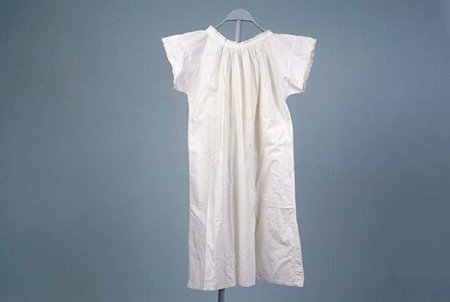 Nattskjorta, möjligen särk, av vitt bomullstyg med mycket korta ärmar. Kantad med uddspets. Knäppes med en knapp i halsen fram. Längd 91 cm
