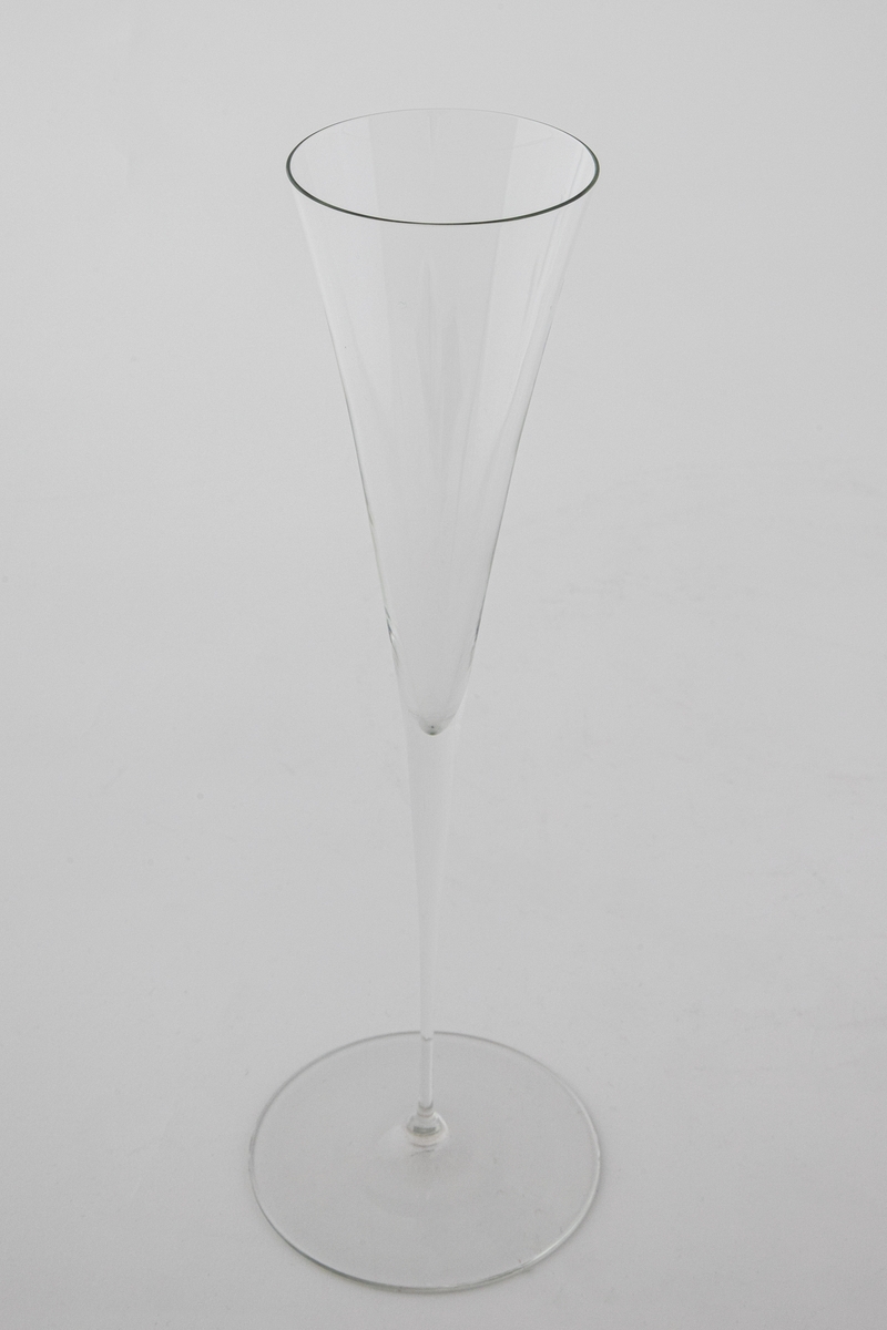Høyreist champangeglass i klart glass. Konisk kupa som glir over i en stett, som hviler på en sirkulær fotplate.