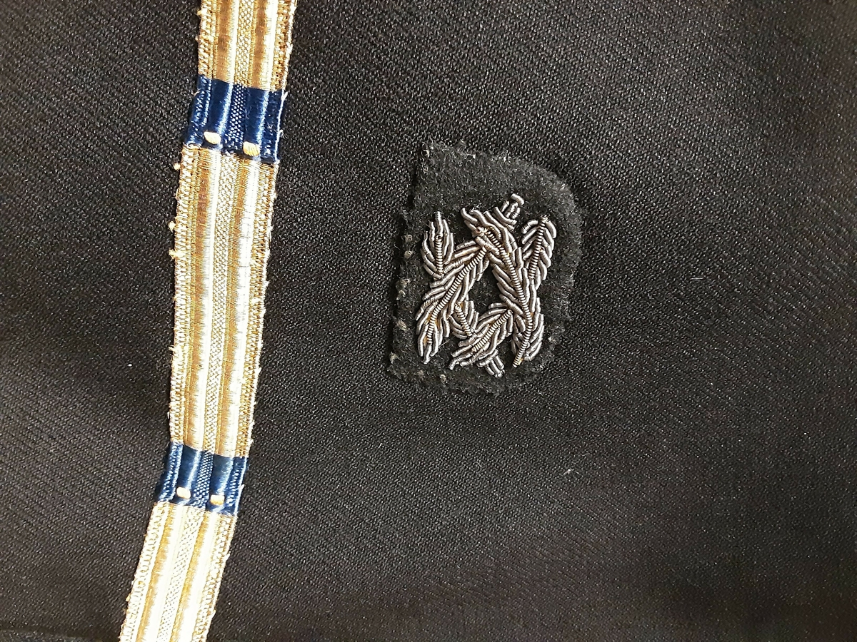 Uniformsjakke tilhørende marineuniform fra 2. verdenskrig. Sort, dobbeltspent med seks gullfargede knapper med ørn i relieff. Nederst på ermet et enkelt gullbånd med blå felter. Over dette båndet påsydd et emblem med små grener. 