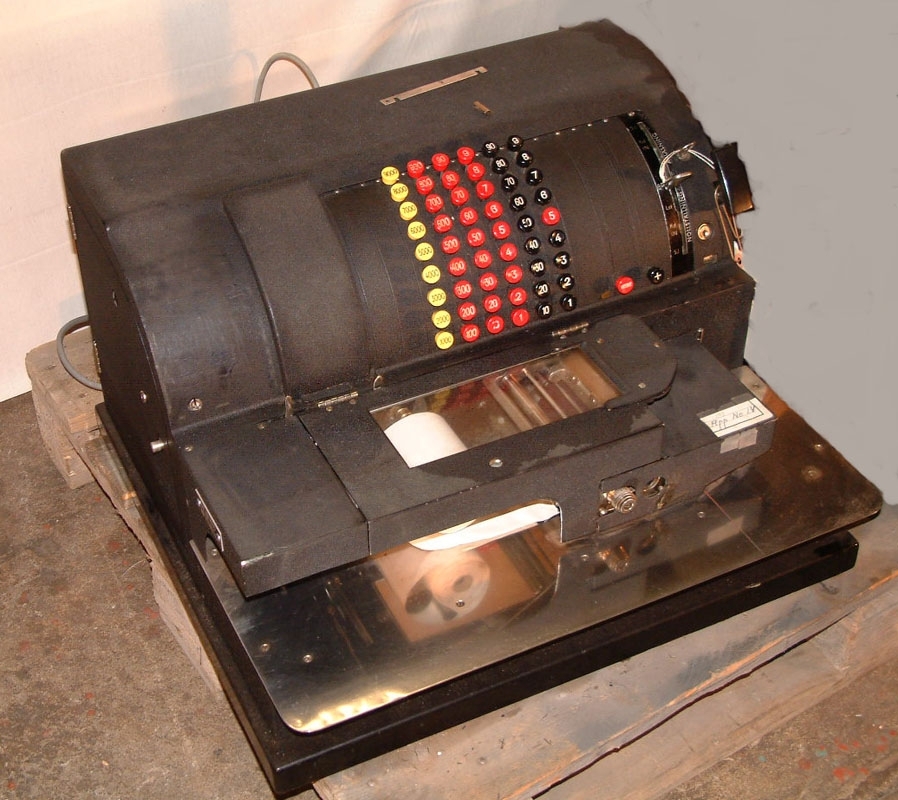 Kassaapparat med svart ytterhölje inom vilket maskineriet sitter. Mitt på den välvda framsidan sitter knappar av röd, gul och svart hårdplast med siffror på, vilka i det här fallet representerar pengasummor. Med knapparna slås siffror in i maskinen som räknar och sedan skriver ut kvitto på papper som kommer ut från en vit pappersrulle.
Längst fram en yta av rostfritt stål lämplig att skriva, lägga papper, fraktsedlar på mm. Med en spak vid sidan om knapparna kan man ställa in "Avläsning" samt "Nollställning". Med en vev i apparatens högra sida matar man fram papper (?). På handskriven fastklistrad lapp står "App No 14". På baksidan en kabel med kontakt som sätts i eluttag för att ge ström till maskinen. 

Genom en gammal adresslapp framkommer att maskinen kommer från Styckegodsterminalen och stationen Stockholm Norra.