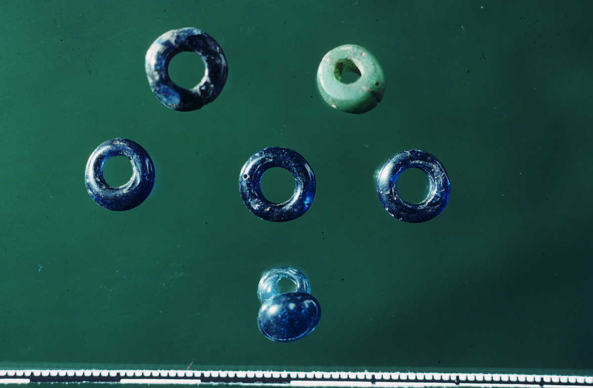 "8 ganske små, ringformete, ensfargete glassperler, 0,3-0,6 cm. Bare fire av dem er hele, alle disse mørkeblå. Dessuten foreligger en grønn perle i to deler som har kunnet settes sammen. Stykkene ble funnet i en meters avstand fra hverandre. Likeså er der et mindre stykke av en lys blå perle, som ble funnet slik. Derimot ble en mørkblå perle og en hvit perle knust under gravningsarbeidet. Her foreligger også en perle eller en liten hengedopp av mørkt blått glass av form nærmest som Müller: Ordning jernalderen fig. 226. 0,8 x 0,9 cm. Den ble funnet liggende igjen i en av de bortkjørte jordhauger fra ifjor, og må rimeligvis opprinnelig ha ligget i en av hustuftene i øst." Ny beskrivelse etter revisjon: Åtte perler av mørk blått, grønt og hvitt glass, herunder fem hele sammenlimte perler og fragmenter av ytterligere tre perler: (1) En sammenlimt, nesten hel avrundet perle med plane, ikke parallelle sider av mørk blått, gjennomskinnelig glass. Stl: 7 mm, diam: 10 mm. (2) Tre avrundede perler uten plane sider av mørk blått, gjennomskinnelig glass. En hel og to nesten hele perler. Stl: 3-4 mm, diam: 9 mm. (3) En sammenlimt og hel avrundet perle med plane, ikke parallelle sider av grønt, opakt glass. Stl: 6 mm, diam: 8 mm. (4) Fjorten fragmenter av minst tre perler, herunder en avrundet perle av hvitt, opakt glass med stl: 6 mm, ytterligere en avrundet mørk blå perle sannsynligvis av form som (1), samt ytterligere en avrundet mørk blå perle av form som (2) med stl: 3 mm.